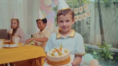 Partide elinde pasta tutan ve kameraya bakan beyaz şapkalı çocuğa odaklan. Arkadaşları arka planda şenlik masasında oturuyor.