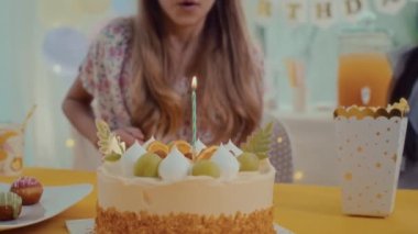 Doğum günü pastasında arkadaşlarıyla masada otururken mumları üfleyen bir kız resmi.