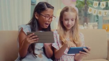 Akıllı telefon ve dijital tablet kullanarak gülen, kanepeye oturan, doğum günü yemekleri ve arka planda süslemeler olan orta boy kızlar.