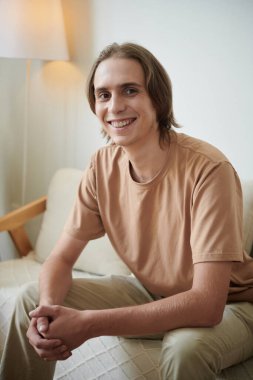 Evdeki koltukta oturan ve kameraya gülümseyen neşeli genç adamın portresi.