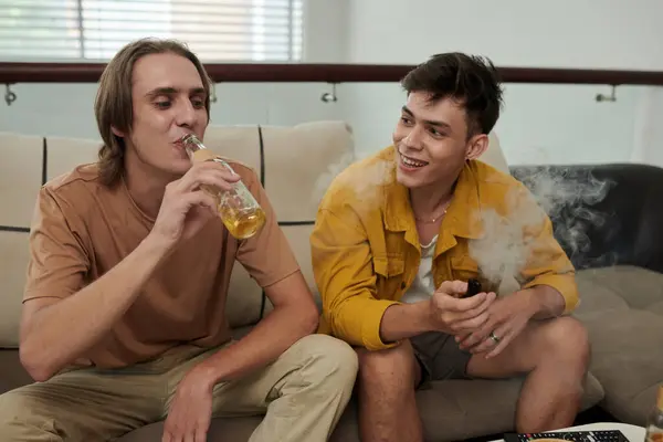 Joyful young people smoking vap and drinking beer when relaxing at home