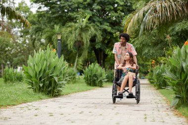 Genç siyahi bir adam, şehir parkında buluşmaktan zevk alırken kız arkadaşının tekerlekli sandalyesini itiyor.