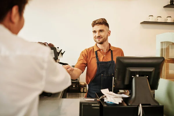 Smiling coffeeshop barista accepting payment for cup of coffee at cashier desk