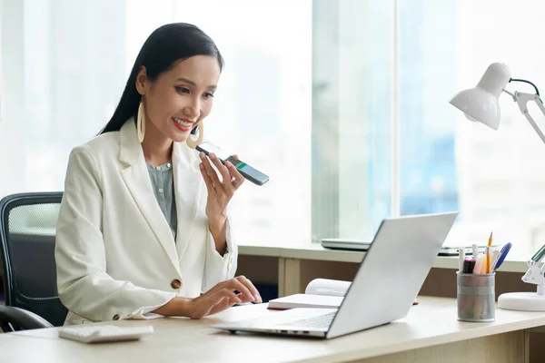 Smiling female entrepreneur checking e-mails on laptop and recording voice messages for colleague