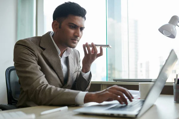 Serious Indian entrepreneur recording voice message for coworker when checking e-mails on laptop