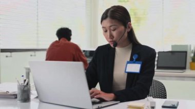 Ofiste dizüstü bilgisayarın başında otururken müşteriyle konuşan çağrı merkezinin pozitif uzman portresi, arka planda iş arkadaşı