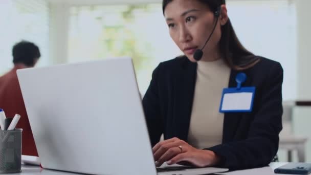 オフィスのラップトップにクライアントにメッセージを入力しながらヘッドセットを着用する若い女性の低角度ビュー — ストック動画