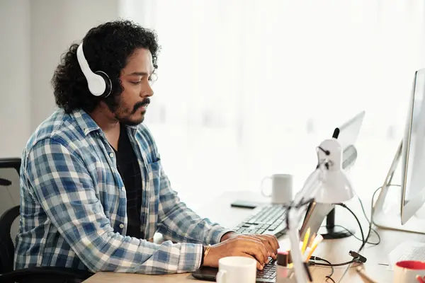 Software developer in plaid shirt listening to music in headphones and coding on laptop