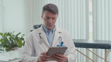 Beyaz önlüklü, elinde tablet olan ve kameraya bakan neşeli sağlık görevlisinin portresi.