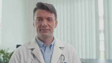 İş yerinde oturan ve kameraya bakan pozitif doktor portresi.
