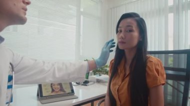 Beyaz önlüklü erkek doktor ve bayan hasta odada oturup dijital tabletteki resme bakarken yüz şekillendirme seçeneklerini tartışıyor.