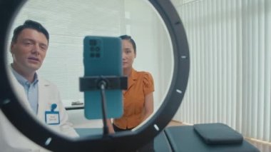Beyaz önlüklü ve tıbbi eldivenli bir erkek doktorun kadın hastasıyla akarsu sırasında kamerayla konuştuğu orta boy bir görüntü.