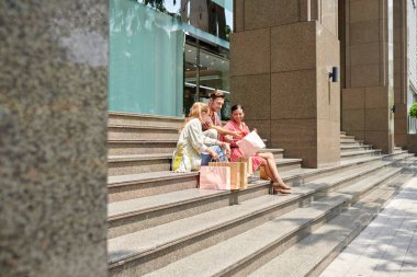 Neşeli arkadaşlar alışveriş merkezinin merdivenlerinde oturup yaptıkları alışverişleri tartışıyor.