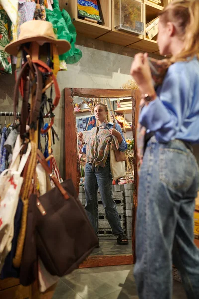 Young woman trying on blouse in front of mirror in thrift shop