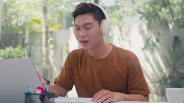 Orta boy kulaklıklı genç Asyalı adamın evde dizüstü bilgisayar kullanarak online ders sırasında notlar alırken görüntüsü.