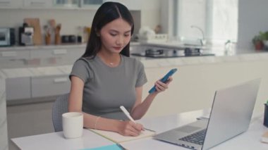 Akıllı telefona bakıp evde ders çalışırken dizüstü bilgisayarın önünde notlar alan konsantre bir kadının orta boy fotoğrafı.