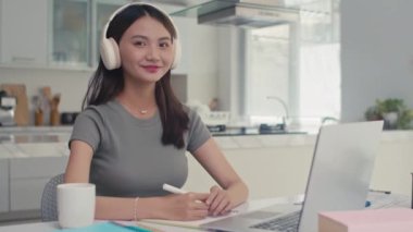 Evde dizüstü bilgisayarın önünde oturan ve kameraya bakan kulaklıklı pozitif kız öğrencinin portresi.