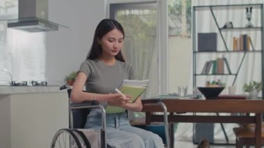 Tekerlekli sandalyede elinde eğitim malzemeleriyle evden ders çalışırken kameraya bakarken orta uzunlukta bir portre resmi.