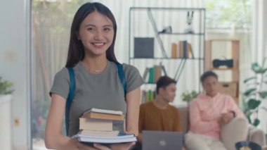 Evde arkadaşlarıyla üniversite projesinde çalışırken elinde kitaplarla gülümseyen Asyalı kızın orta boy portresi.