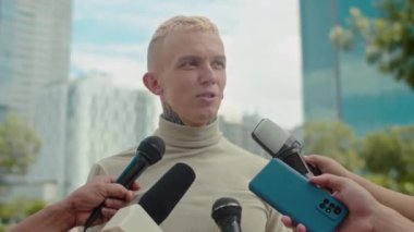 Gazetecilerin sorularını dışarıda mikrofonlara yanıtlayan boynunda dövmesi olan genç bir adamın orta boy fotoğrafı.