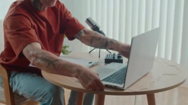 Erkek podcaster stüdyoda kayıt yapmadan önce laptop ve mikrofonla masada otururken ekipman hazırlıyor.