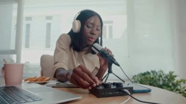 Orta boy Afrika kökenli Amerikalı kadın podcast sunucusu kulaklık takarken stüdyoda masada oturan sesleri kontrol ederken.