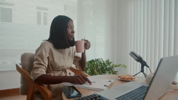 アフリカ系アメリカ人ジャーナリストがコーヒーを飲んでスタジオに座っている彼女のポッドキャストの脚本について考える中長いショット — ストック動画
