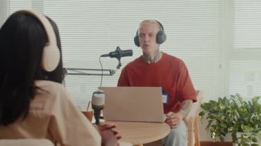 Kulaklıklı kadın konuğun omzundan yayına giren erkek sunucuyla stüdyoda dizüstü bilgisayar ve mikrofon kullanarak konuşuyor.