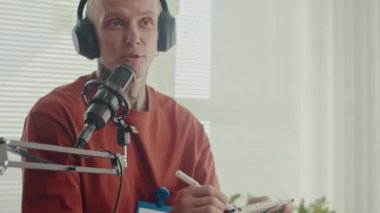 Orta boy beyaz erkek sohbet programı sunucusu kulaklıklar takıyor ve stüdyodaki konukla röportaj yaparken mikrofona konuşuyor.