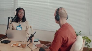 Orta boy, kulaklıklı Afrikalı Amerikalı gazetecinin stüdyosundaki erkek girişimciyle röportaj yaparken laptopun önünde oturması.
