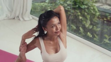 Eğilerek neşeli Asyalı kadının yoga yaparken bacak bacak üstüne atıp minder üzerinde antrenman yaptığı sahneyi çek.