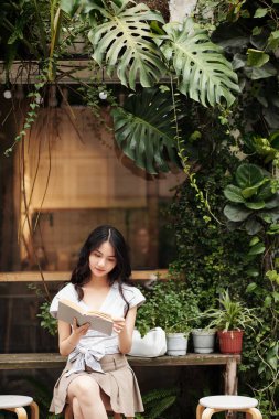 Genç Asyalı kadın otantik kafenin dışında oturmuş kitap okuyor.