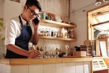 Kahveci garson müşteriyle telefonda konuşuyor ve sipariş detaylarını yazıyor.