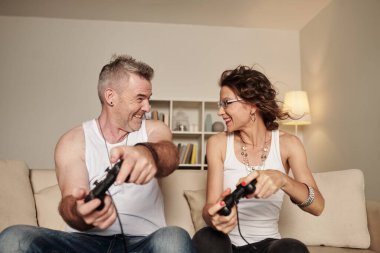 Kolsuz tişörtlü, video oyunu oynayan ve birbirlerine bakan orta yaşlı bir çift.