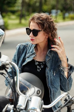 Güneş gözlüklü göz kamaştırıcı olgun kadın motosiklette oturuyor ve başka tarafa bakıyor.