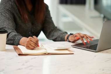 Diz üstü bilgisayar ekranından raporları kontrol eden ve planlamacıdan not alan kadın girişimcinin kırpılmış görüntüsü