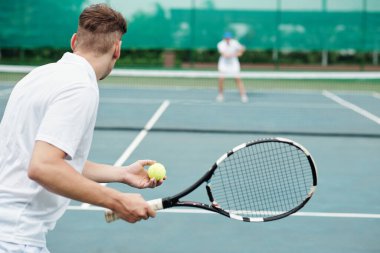 Young man playing tennis with friend and serving ball, view from back