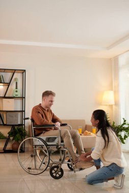 Umursayan kız arkadaş tekerlekli sandalyede oturan arkadaşının ayakkabı bağcığını bağlıyor.