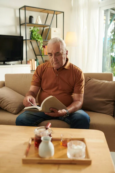 Starší Muž Pije Čaj Čte Podmanivou Knihu Doma — Stock fotografie