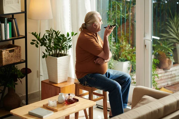 Lonely senior man looking outside through glass doors and recording voice message for friend