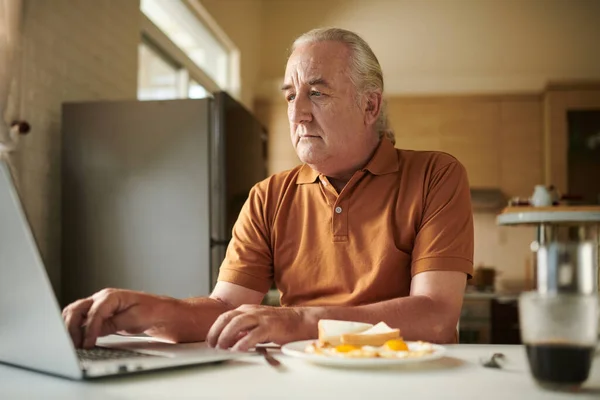 Seriös Senior Man Äter Frukost Och Arbetar Laptop Vid Köksbänken — Stockfoto