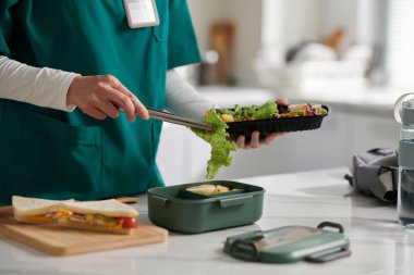 Tıp hemşiresi iş yerinde öğle yemeği için plastik kabın içinde salata ve sandviç paketliyor.