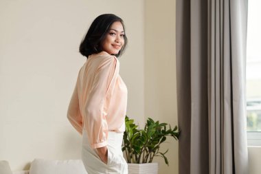 Açık şeftali rengi ipek bluzlu zarif Asyalı kadın odada dikiliyor ve kameraya doğru dönüyor.