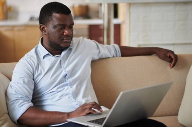 Evdeki kanepede oturan siyah adam dizüstü bilgisayarda çalışıyor, e-postaları cevaplıyor ya da belgeleri kontrol ediyor.