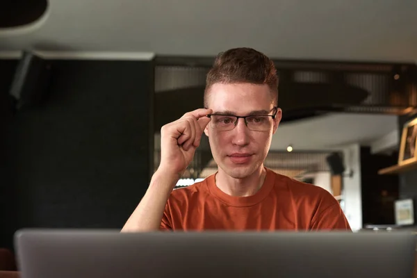 Smiling young man adjusting glasses when reading e-mails on laptop or looking for bugs