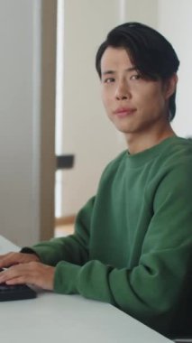 Bilgisayarda çalışan ve ofisteki kameraya bakan neşeli erkek kodlayıcının dikey görüntüsü