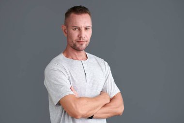 Gri tişörtlü, kollarını kavuşturup kameraya bakan kendinden emin olgun bir adamın portresi.