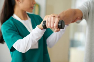 Rehabilitasyon merkezi çalışanının kontrolü altında küçük bir dambılla egzersiz yapan adam.