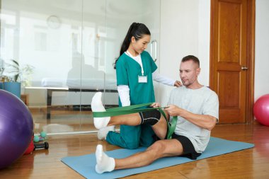 Rehabilitasyon merkezinin kontrolü altında direniş bandıyla egzersiz yapan olgun bir adam.