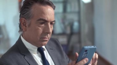 Ofiste çalışırken ve kameraya bakarken akıllı telefon kullanan yaşlı finans müdürünün portresini kapat